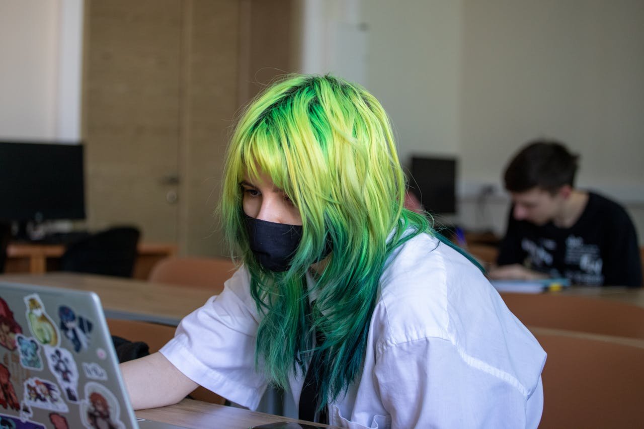 High school student with dyed green hair uses a laptop wearing a mask in classroom setting.