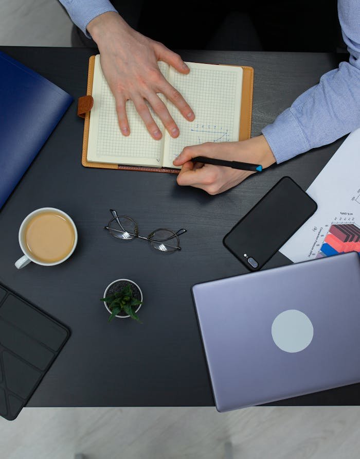 Overhead view of a workspace with a notebook, coffee, and electronic devices.