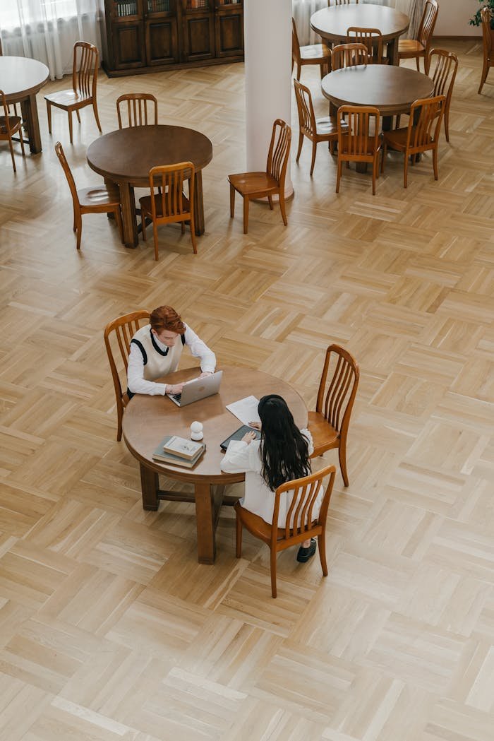 Two students at a wooden table using a laptop in a quiet library setting.